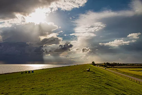 Deichlandschaft mit dramatischem Himmel und Sonnenstrahl über der Nordsee auf Pellworm